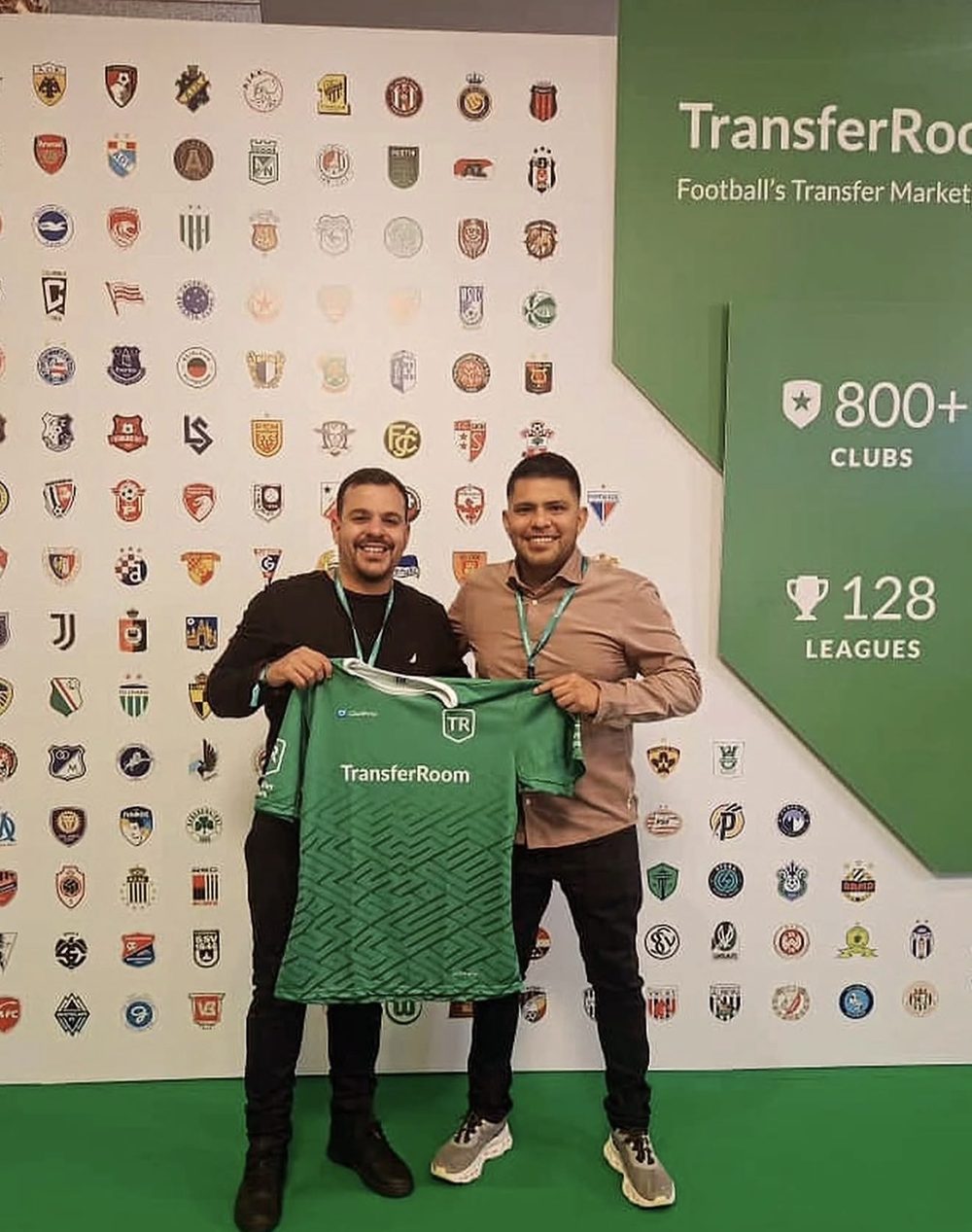 Two men holding up a green TransferRoom shirt in front of a wall of club crests