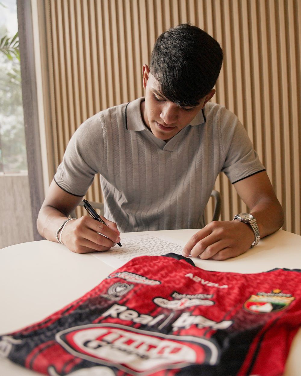 Un jugador joven firma un contrato sobre una mesa, con una camiseta roja y negra del Deportivo Lara en primer plano